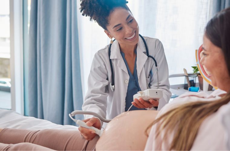 A pregnant woman is at the clinic getting her sonogram as she excitedly smiles at her provider who is holding a medical device on her stomach.