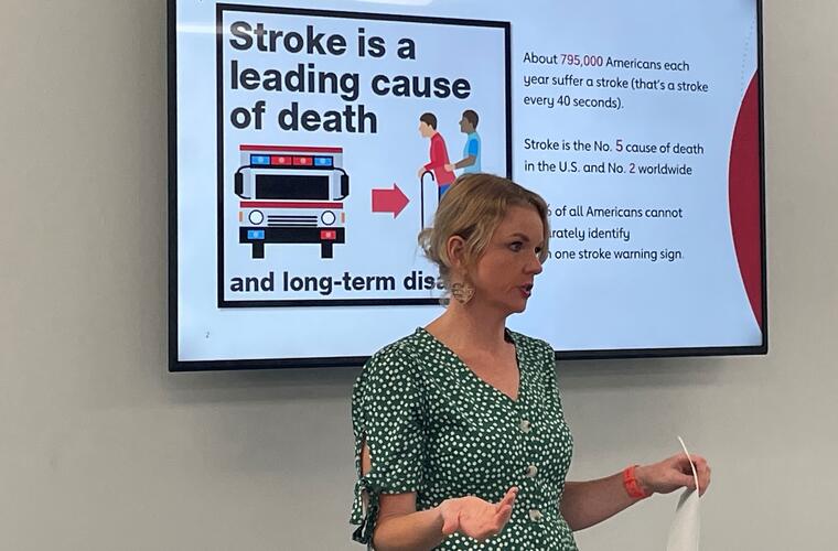 A woman with short blonde hair pulled back wearing a green dotted dress stands in front of a wall-mounted TV screen. The text on the screen behind her reads: Stroke is a leading cause of death and long-term disability.  The right side of the screen has the text that reads:  About 795,000 Americans each years suffer a stroke (that's a stroke every 40 seconds).  Stroke is the No. 5 cause of death in the U.S. and No. 2 worldwide.
