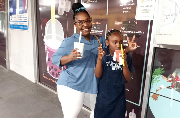 Black woman in a blue top and white pants stands beside a young Black girl wearing blue overalls both are enjoying a treat.