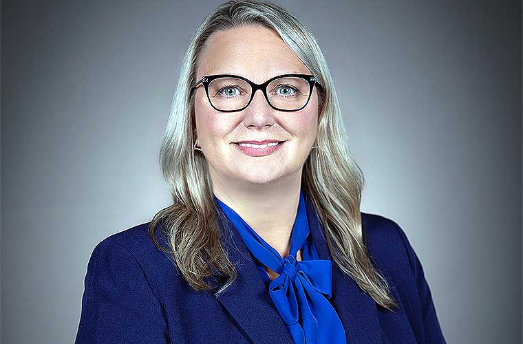 A professional headshot of Lynette Clinton smiling. She is wearing glasses, a navy jacket, a blue shirt with a bow tied in the front and she has wavy dark blonde hair.