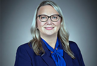 A professional headshot of Lynnette Clinton smiling. She is wearing glasses, a navy jacket, a blue shirt with a bow tied in the front and she has wavy dark blonde hair.