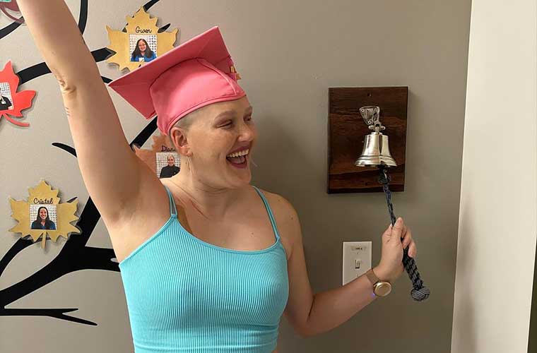 A woman wearing a pink graduation cap celebrates and smiles as she rings a bell which signifies the end of cancer treatment.