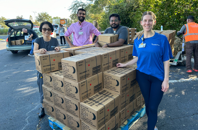 At a food distribution event, two men and two women stand next to a pallet stacked with boxes.