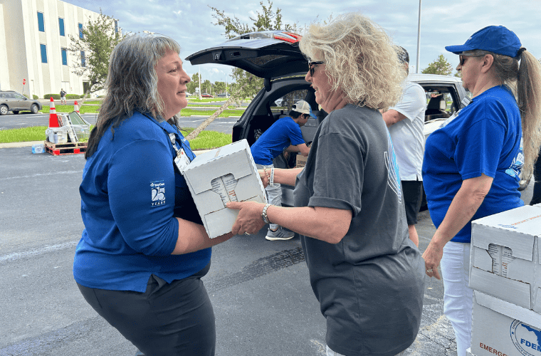 At a food distribution event, three women in blue and grey shirts are delivering boxes to drivers waiting in line. 