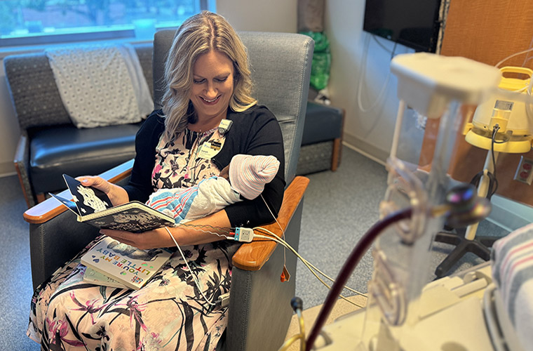 A woman sitting in a chair is holding a premature baby who is wearing a pink hat. The woman is reading a book to the baby.