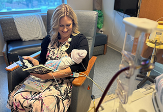 A woman sitting in a chair is holding a premature baby who is wearing a pink hat. The woman is reading a book to the baby.