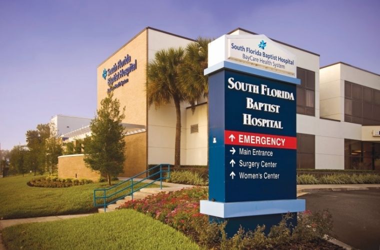 A clear view of a white and blue sign displays South Florida Baptist Hospital and the hospital's logo against a bright sky.