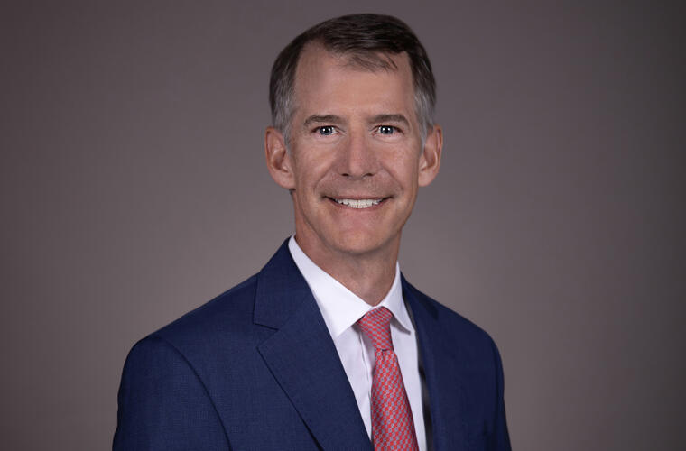 BayCare Medical Group Vice President and Chief Medical Officer Dr. Timothy Liesching, waist-up shot, older white male, short graying hair, smiling, dark blue blazer, red dotted tie, white shirt, gray gradient background.