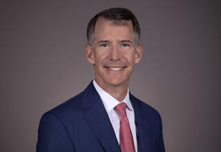 BayCare Medical Group Vice President and Chief Medical Officer Dr. Timothy Liesching, waist-up shot, older white male, short graying hair, smiling, dark blue blazer, red dotted tie, white shirt, gray gradient background.