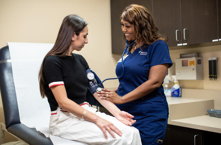 A BayCare team member in navy blue scrubs measures the blood pressure of a seated patient in a clinical setting, using a cuff and stethoscope while medical supplies are visible in the background.