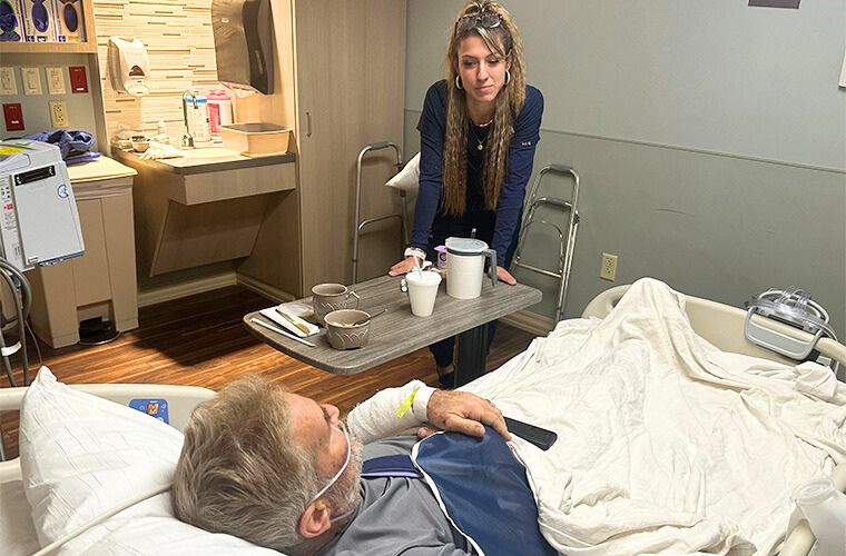 Woman in blue scrubs stands over a hospital bed with a patient.