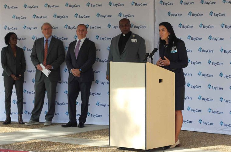 A woman in a navy blue pant suit stands behind a podium with BayCare in the background. Next to her stand three men and a women dressed in business causal.