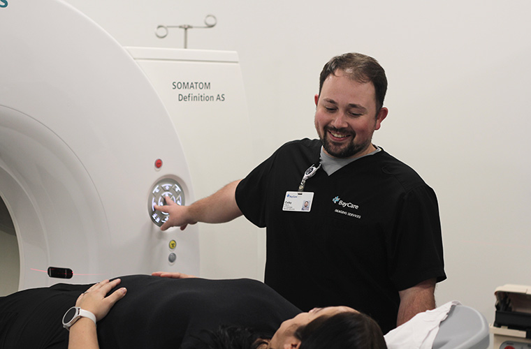 A man wearing black scrubs with the word "BayCare" on them smiles, as he looks down and uses his right hand to press buttons on a control panel of a CT scanning unit in a clinical room. A woman is lying on the CT unit's bed and looking up at him.