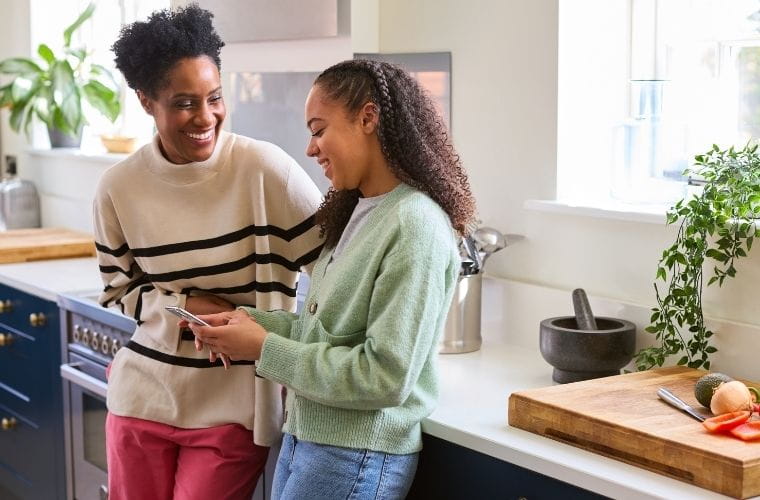 A mother talks with her teenage daughter as they stand in a kitchen smiling at each other over a smartphone, sharing something on the screen.