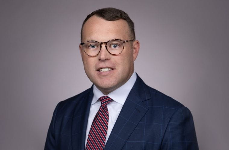 A formal smiling headshot of Chase Hall wearing a dark blue checkered suit jacket, a white dress shirt and a red-and-navy striped tie, posed against a plain gray background.