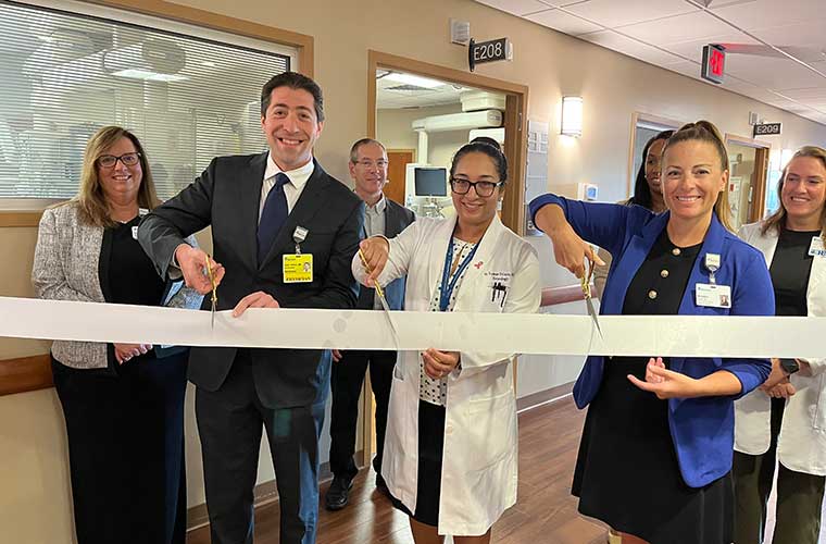 A group of men and women, some dressed in hospital scrubs, surround three people holding scissors as they prepare to cut a ribbon to open a unit in a hospital.