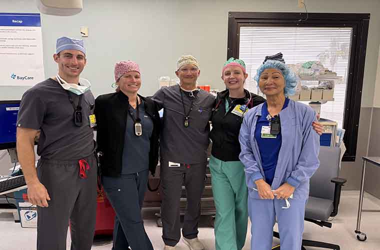 Five medical professionals wearing scrubs and surgical caps, smiling in a hospital room with a computer and medical equipment in the background. The BayCare logo is visible on a sign.