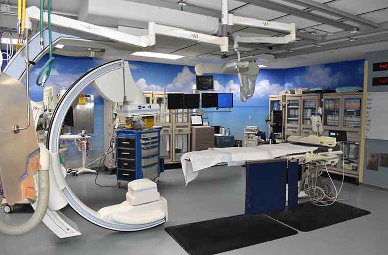 A modern medical imaging room with an advanced X-ray imaging system, a hospital bed, and various medical equipment and monitors. The room is well-lit and spacious.