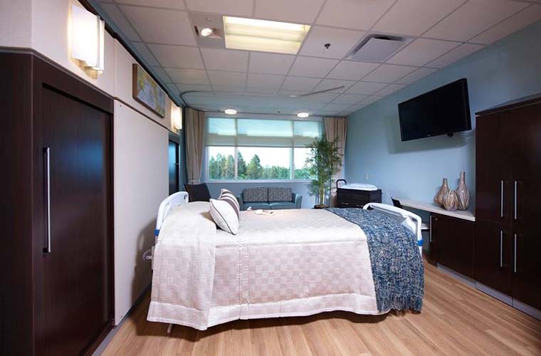 A hospital room featuring a single bed covered with a white and blue blanket, a, baby crib, a flat-screen TV on the wall, and a large window showing a view of trees.