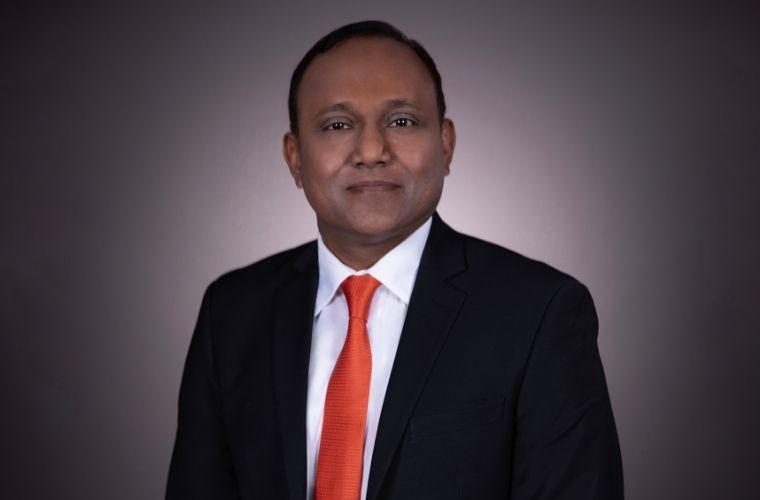 A smiling headshot of Dr. Shelby Kutty. He is wearing a black suit jacket, a white button shirt and a red tie.