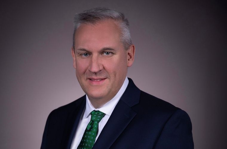 A headshot of Marc Hallee smiling, wearing a a formal black suit jacket, white button-up shirt and green tie.