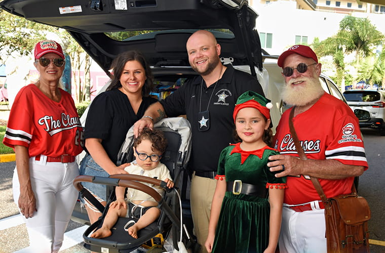 Six people pose together in front of a vehicle trunk, including a child in a stroller, a police officer, two people in "The Clauses" shirts, and a girl dressed as an elf.