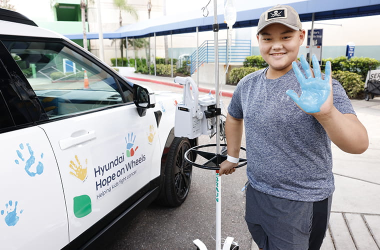 A boy with blue paint on his hand and holding an IV pole stands in front of white vehicle with colorful handprints.