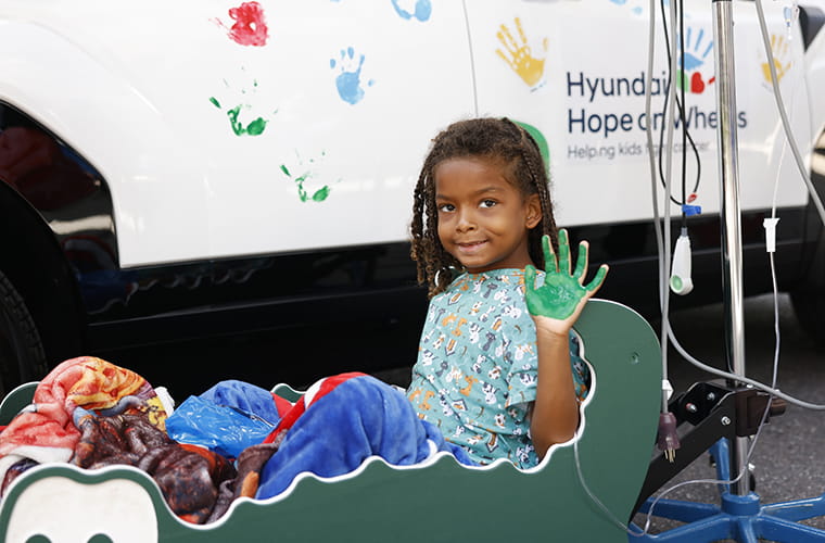A young boy in a hospital wagon holds up hand with green paint.
