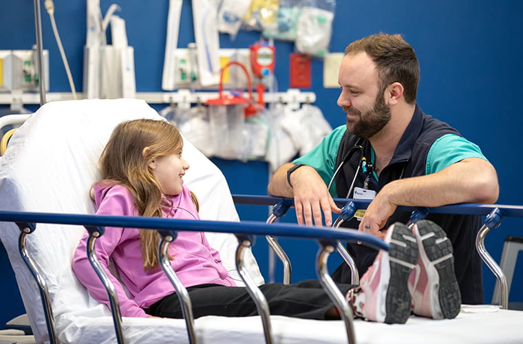 Young girl in pink sweater laying on hospital bed talking to a male physician kneeling by her bed.