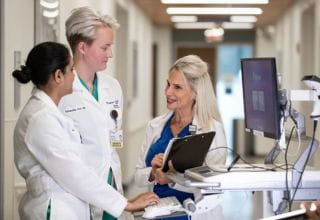 Three women in white lab coats stand together in a hospital hallway engaged in conversation.