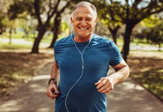 An older man wearing athletic clothing and headphones, jogging in a park surrounded by greenery and trees.