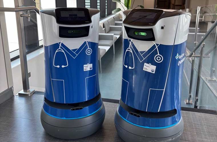 Two small blue and white robots decorated in health care scrubs stand together in the hallway of a building.