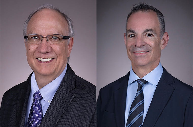 Two men are wearing blue shirts, ties and dark suits. One man is wearing eyeglasses.