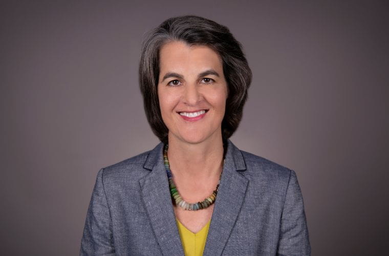 A smiling headshot of Keri Eisenbeis. She is wearing a gray jacket with a yellow V neck shirt underneath and a statement necklace made up of large, cylindrical beads in a range of earthy tones. She has short, straight graying hair.