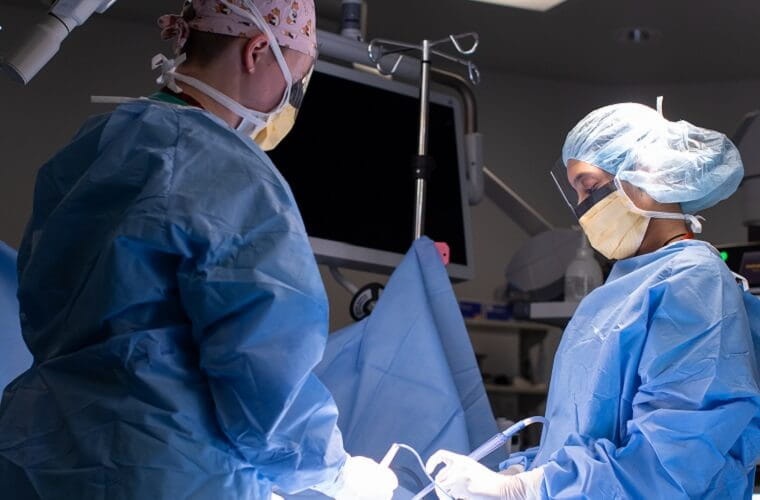 Two physicians are wearing masks and scrubs while performing a procedure in a hospital surgical suite.