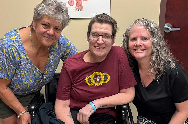 A woman wearing glasses and a maroon T-shirt with sunflowers on it sits in a wheelchair and is flanked by two other women.