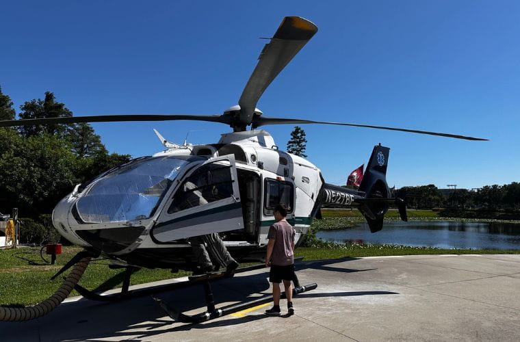 A young man stands beside a parked helicopter on the ground with a lake in the background on the campus of St. Joseph's Hospital.