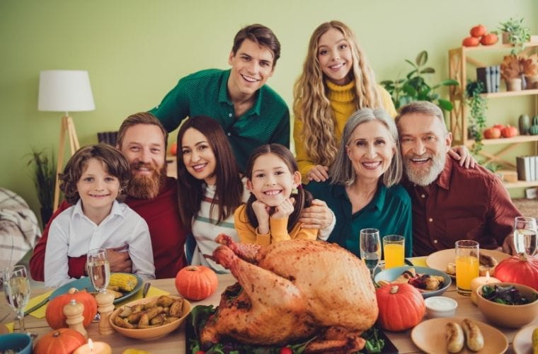 A festive dining table set for a holiday meal, featuring a large roasted turkey as the centerpiece surrounded by pumpkins, vegetables, and assorted dishes. Glasses of orange juice and champagne are placed on the table. In the background, a group of people are gathered closely, with shelves decorated with plants and seasonal items behind them.