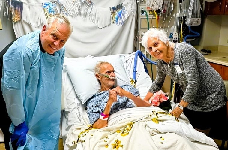 A hospital patient, Anthony, is lying in a bed with white sheets decorated with gold ribbon pieces, wearing a patterned gown and a pink wristband. The patient is holding hands with a visitor, Nancy, standing on the right, who is leaning slightly forward, smiling and holding a bouquet of pink flowers. Another person on the left, Father John, is wearing a full blue protective gown and gloves, standing close to the bedside. Behind the bed, silver and white fringe garlands are draped as festive decorations. The background includes medical equipment, tubing, and cabinets, with a white balloon visible near the upper right corner.