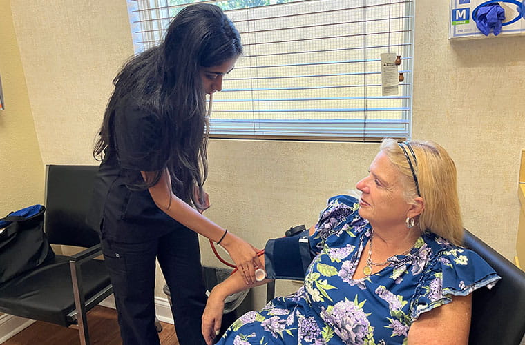 A woman with long dark hair wearing black medical scrubs uses a stethoscope and blood pressure cuff to take the blood pressure of a woman with blond hair wearing a flower print dress who is sitting in a chair at a doctor's office.