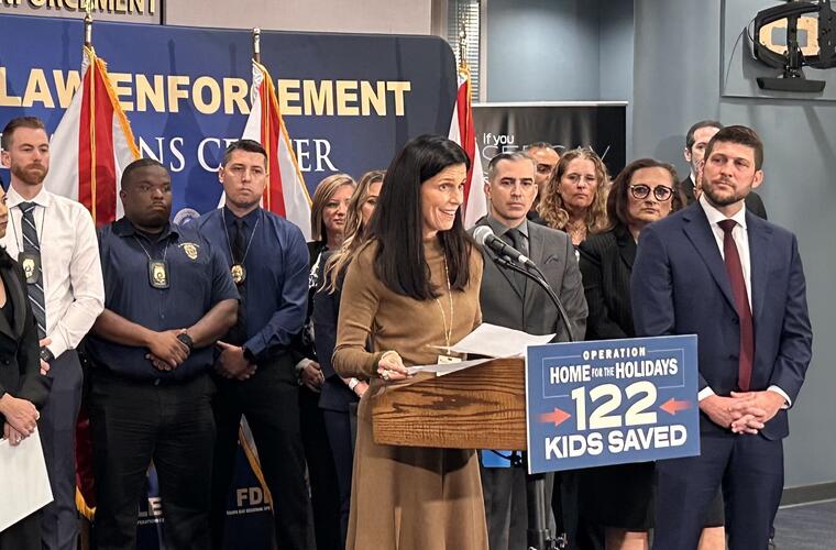 BayCare Behavioral Director of Operations Tracey Kaly speaking at a podium during a press conference, surrounded by law enforcement officials and a banner featuring the text 'Operation Home for the Holidays: 122 Kids Saved'.