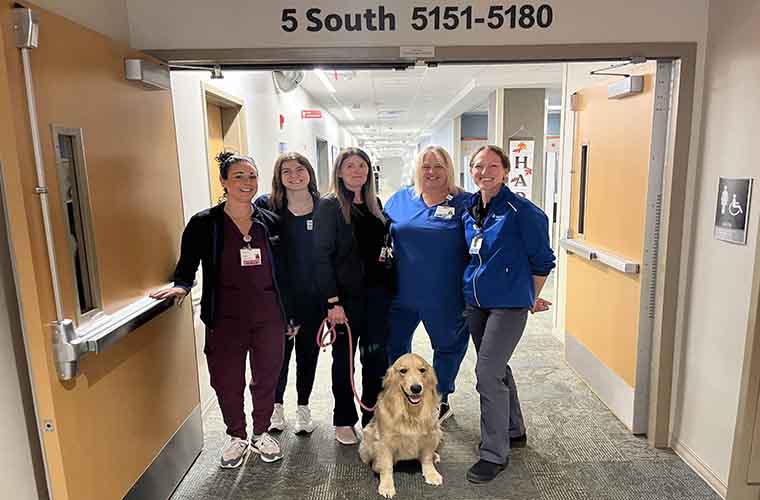 Five health care professionals and a service dog standing in a hospital corridor, smiling at the camera.