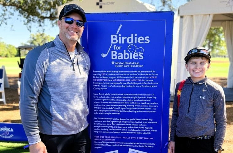 A man and a young boy stand smiling beside a large “Birdies for Babies” sign at the ANNIKA driven by Gainbridge at Pelican event.