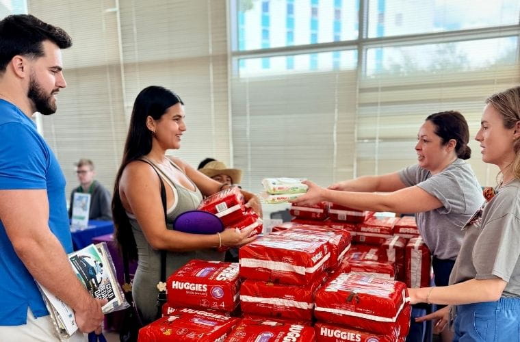 Two event volunteers hand a pregnant mother, who is with family, diapers and baby wipes across a table stacked with Huggies packages at the BayCare Kids Family Expo.