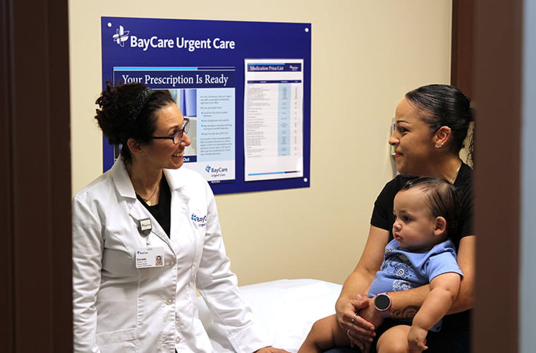 A physician assistant wearing a white medical coat and a BayCare badge speaks with a mother and toddler-aged son in an examination room. Signage on the wall behind them reads "BayCare Urgent Care" and "Your Prescription Is Ready."