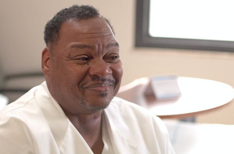 A man wearing a white shirt smiles warmly while seated indoors near a window, with soft natural light illuminating his face.