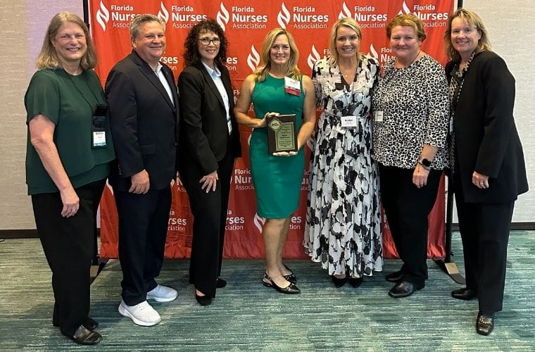 A group of people pose with a plaque. A woman in green in the middle of the group is holding the award.