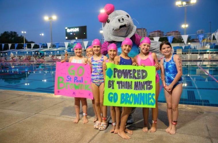 A group of young swimmers in pink caps pose poolside with a manatee mascot, holding bright signs that read “Go St. Paul Brownies” and “Powered by Thin Mints, Go Brownies.”