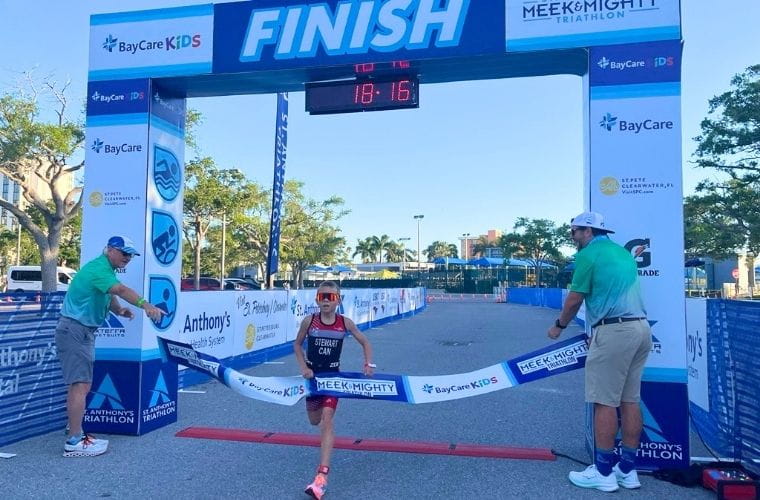 A young athlete wins a kids triathlon, breaking the finish-line tape under the race arch.