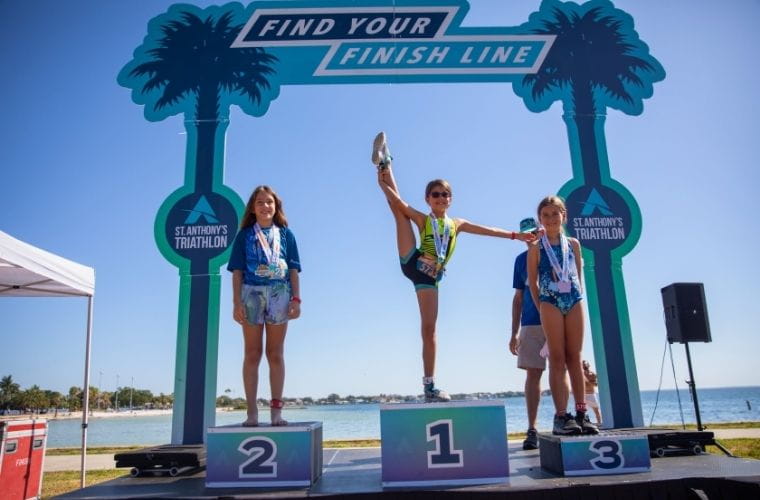 Three young girls stand on a winners podium at a triathlon, medals around their necks, with the first-place winner striking a celebratory pose.